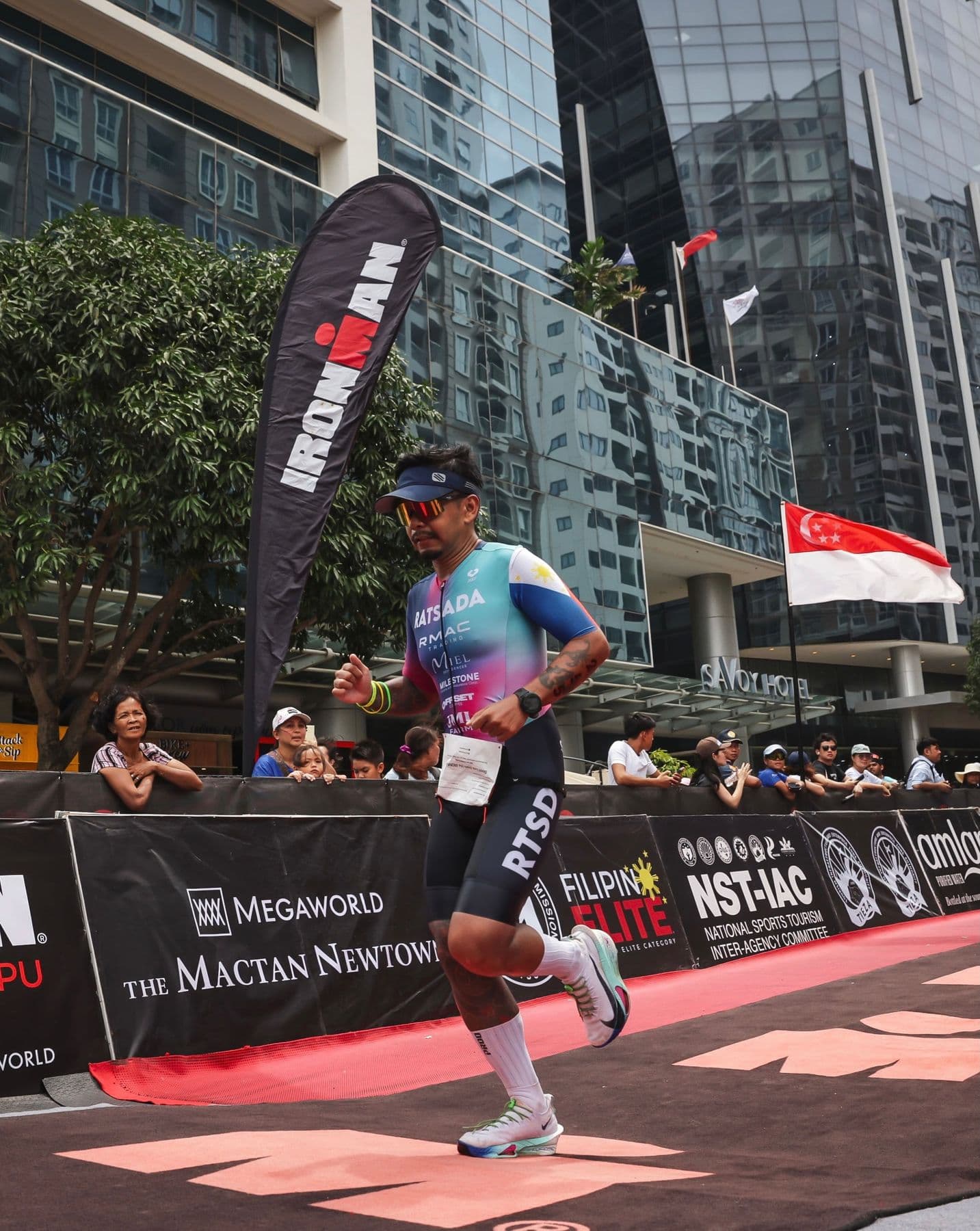 Triathlete running down a finishing chute past an IRONMAN flag between city towers